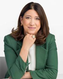 A mature woman wearing a green blazer sits at a desk with her hand under her chin looking confident and relaxed