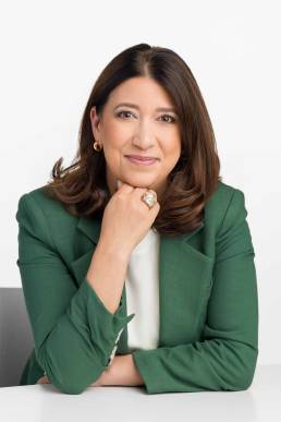 A mature woman wearing a green blazer sits at a desk with her hand under her chin looking confident and relaxed
