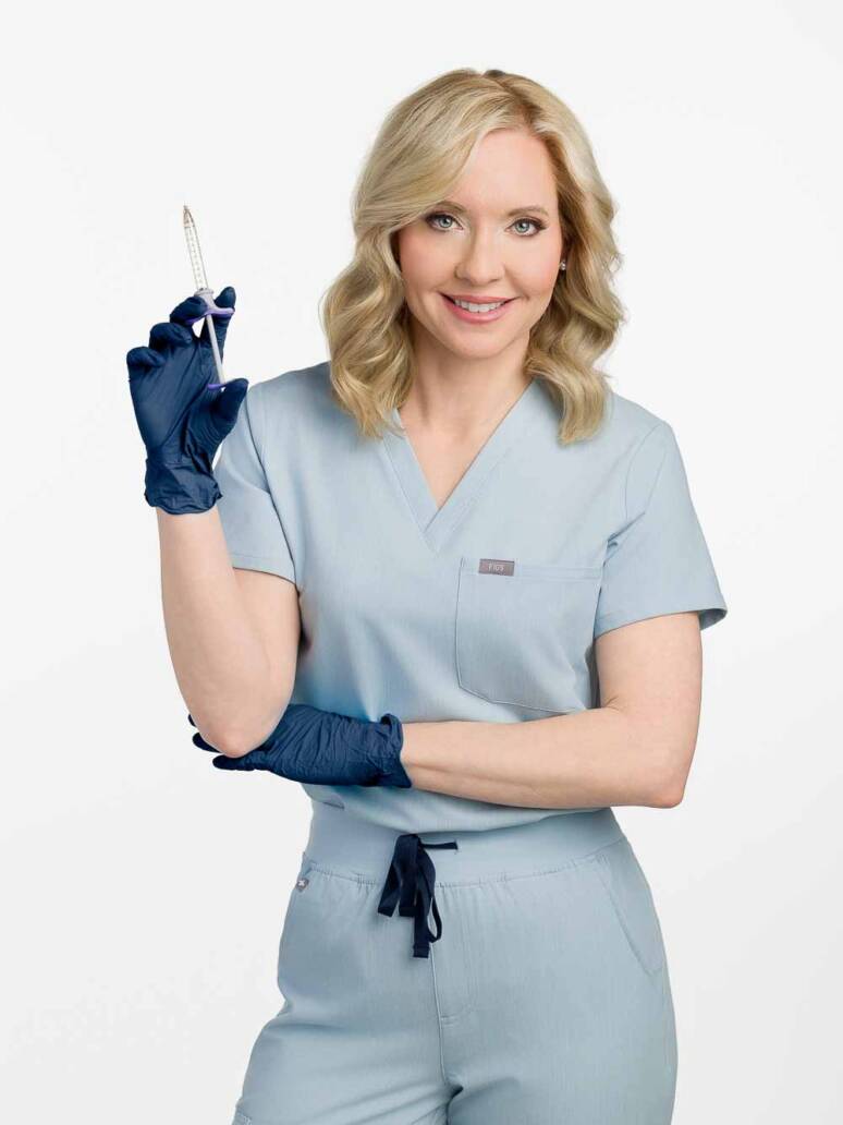 An medical injector wearing gloves holding a needle and smiling warmly.