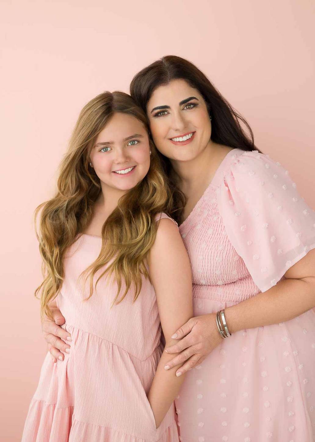 A portrait of a mother and grade school age daughter wearing light pink dresses with a light pink background