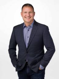 A man in a sport coat and jeans stands with his hands in his pants pockets looking friendly and approachable