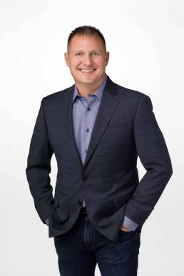 A man in a sport coat and jeans stands with his hands in his pants pockets looking friendly and approachable