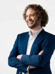 A man wearing a blue suit and white button-up dress shirt with curly hair standing looking to the side with a big welcoming expression on his face.