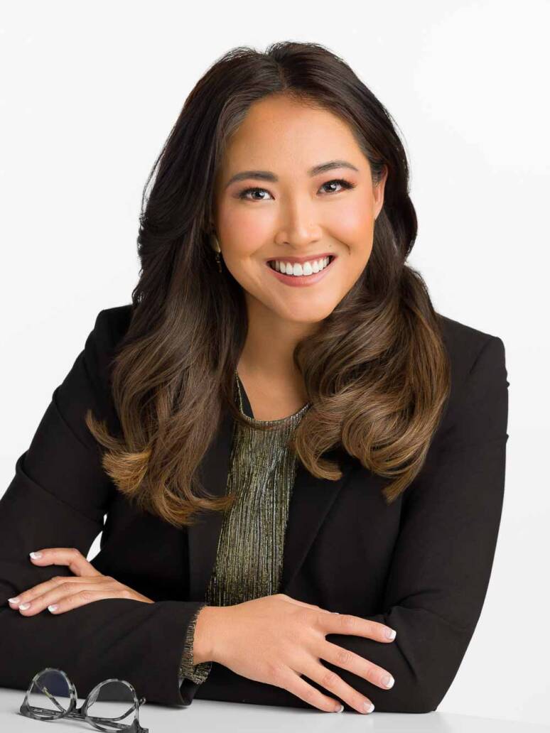 A female psychiatrist leaning forward on a white table with her arms folded and smiling at the camera looking knowledgable and approachable.