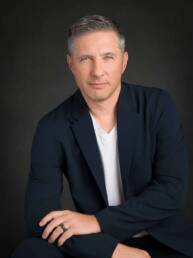A handsome portrait of an executive man in a blazer and tshirt sits casually with a serious but approachable expression.
