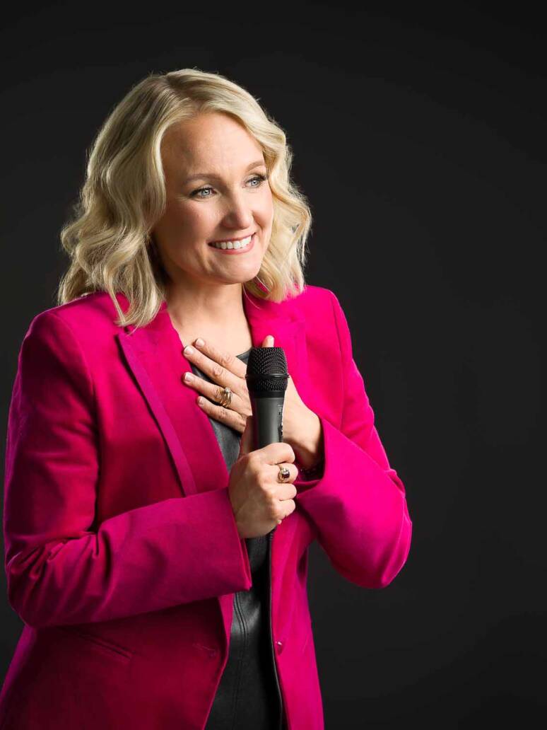 A female speaker standing in the spotlight wearing a magenta blazer and holding a mic looking to the side with her hand on her chest in gratitude.