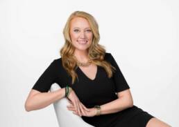 A beautiful blonde Arvada woman sitting confidently in a sideways office chair wearing a black bodycon dress.