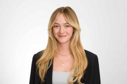 A corporate headshot of a young blonde female smiling at the camera looking proud and happy wearing a black blazer.