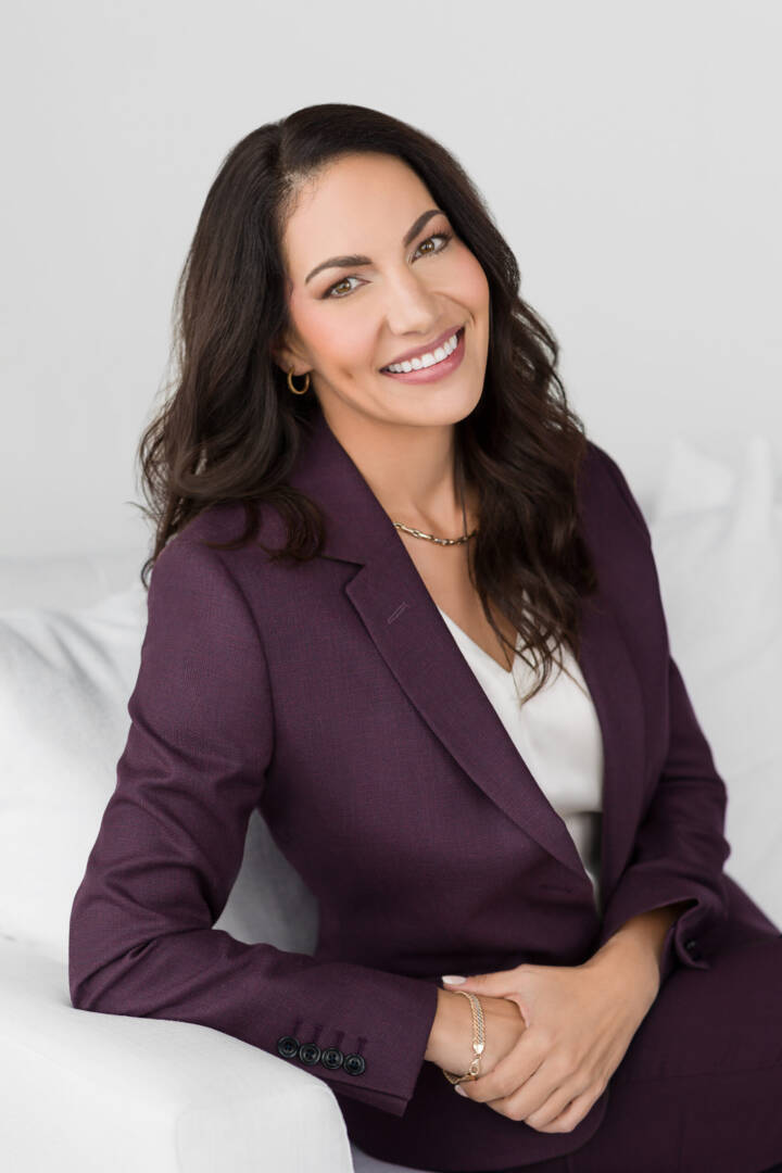 A brunette woman with long hair wearing a dark purple suit sitting on a white sofa looking helpful and friendly and smiling at the camera.