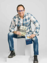 A full body personal branding photo of a man sitting on boxes leaning forward holding two books he authored.