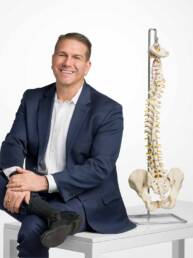 A male chiropractor in a blue suit sits on a table next to a prop spine smiling looking confident, friendly, and knowledgable.