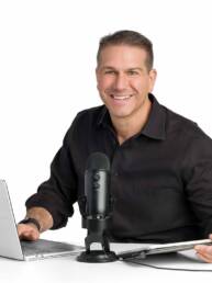 A mid-life man sits at a desk with his laptop and podcast mic smiling at the camera as he's about to record live.