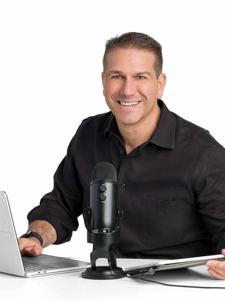 A mid-life man sits at a desk with his laptop and podcast mic smiling at the camera as he's about to record live.