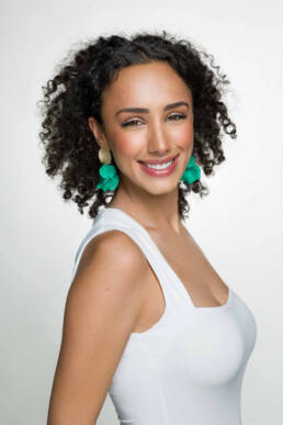 A 20-something woman wearing a white tank top and big earrings standing angled smiling at the camera.
