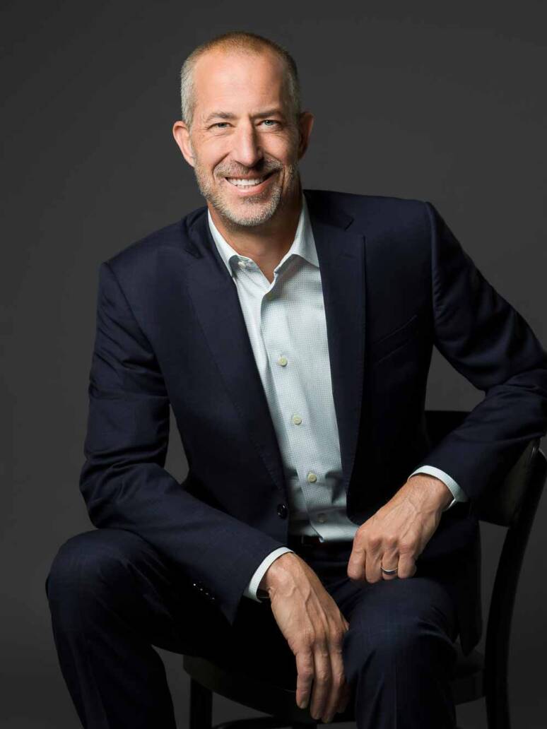 A Denver attorney wearing a navy suit sits casually on a chair while leaning on his forearms and looking approachable and welcoming.