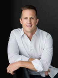 A Denver man wearing a white button-up with the sleeves rolled up sitting casually on a chair looking serious, but casual.