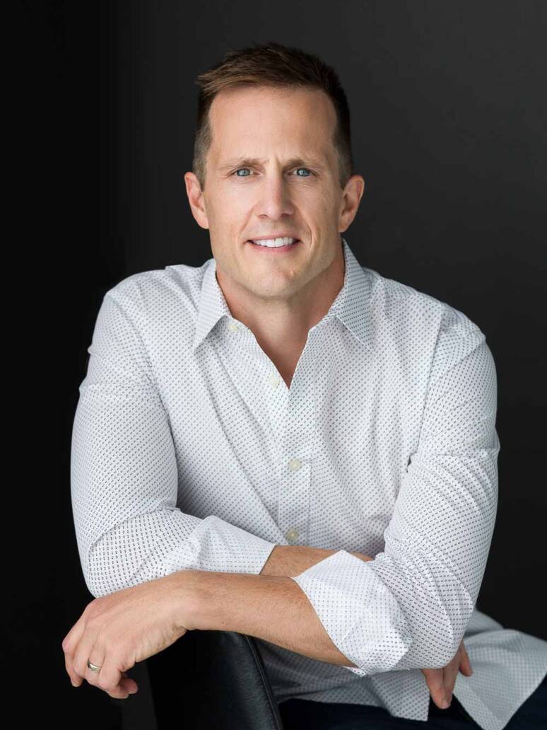 A Denver man wearing a white button-up with the sleeves rolled up sitting casually on a chair looking serious, but casual.