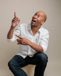 A man with an excited smile wearing jeans and a white button-up sitting casually and using hand gestures to point to the side.