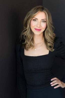 A woman leaning on a black wall with her hand on her waist smiling peacefully at the camera.