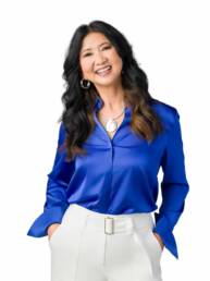 A woman wearing white slacks and a royal blue blouse stands casually with her hands in her pockets and head tilted looking very friendly.