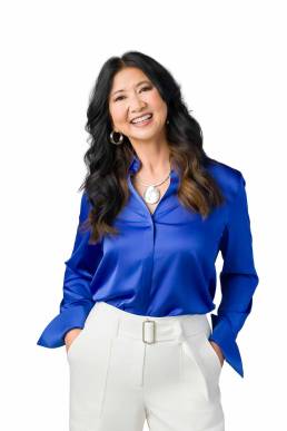 A woman wearing white slacks and a royal blue blouse stands casually with her hands in her pockets and head tilted looking very friendly.