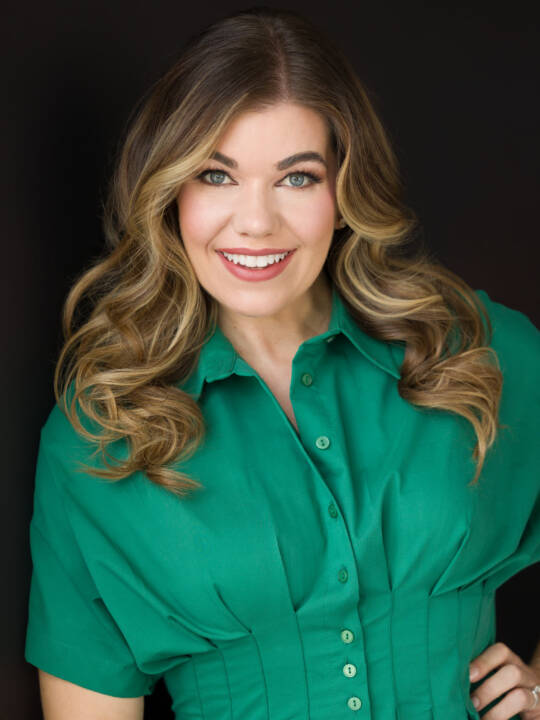 A female in a green dress leaning on a black wall smiling at the camera photographed by award-winning headshot photographer Rachel Sorbet