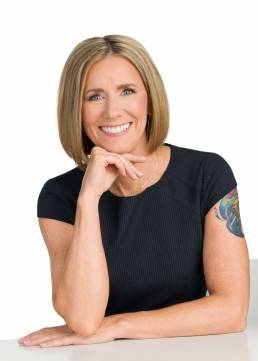 A blonde woman with a bob haircut sits at a white table smiling at the camera with her hand under her chin