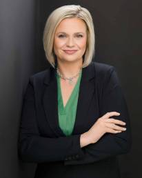 A blonde executive wearing a black suit standing leaning on a wall with her arms folded and smiling confidently
