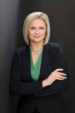 A blonde executive wearing a black suit standing leaning on a wall with her arms folded and smiling confidently