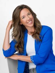 A long haired woman sits casually in an office chair wearing a royal blue blazer, white shirt and jeans