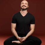 A man sitting barefoot on the floor in black yoga clothes smiling with inner peace and joy