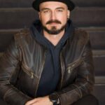 A man wearing a leather jacket and fedora sitting on a staircase looking serious and direct