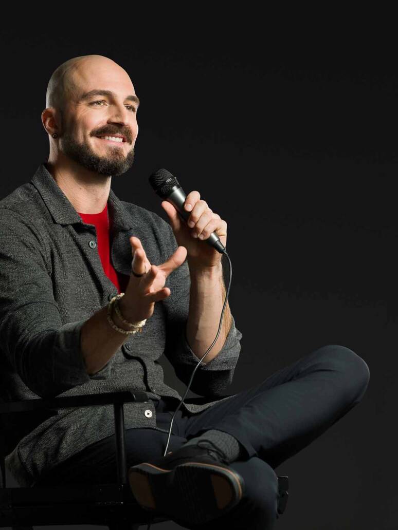 A smiling man sitting in a director's chair on stage holding a mic and speaking with hand gestures