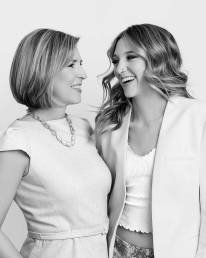 A mom and adult daughter with their back arms around each other looking at each other and smiling