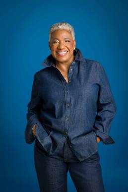 A middle-life woman standing confidently with a big smile on her face and her hands in her jeans pockets on a blue backdrop